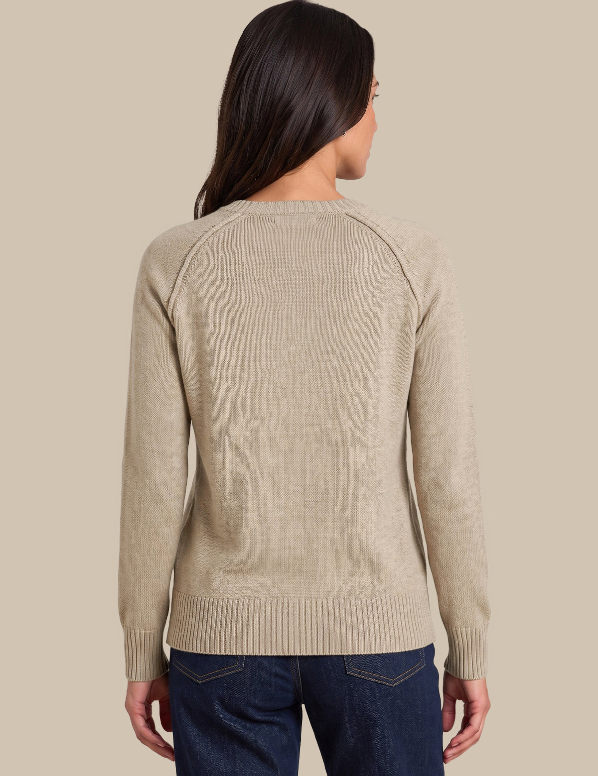 A woman viewed from the back wearing a light beige knit sweater with raglan sleeves and dark blue jeans, set against a solid beige background.
