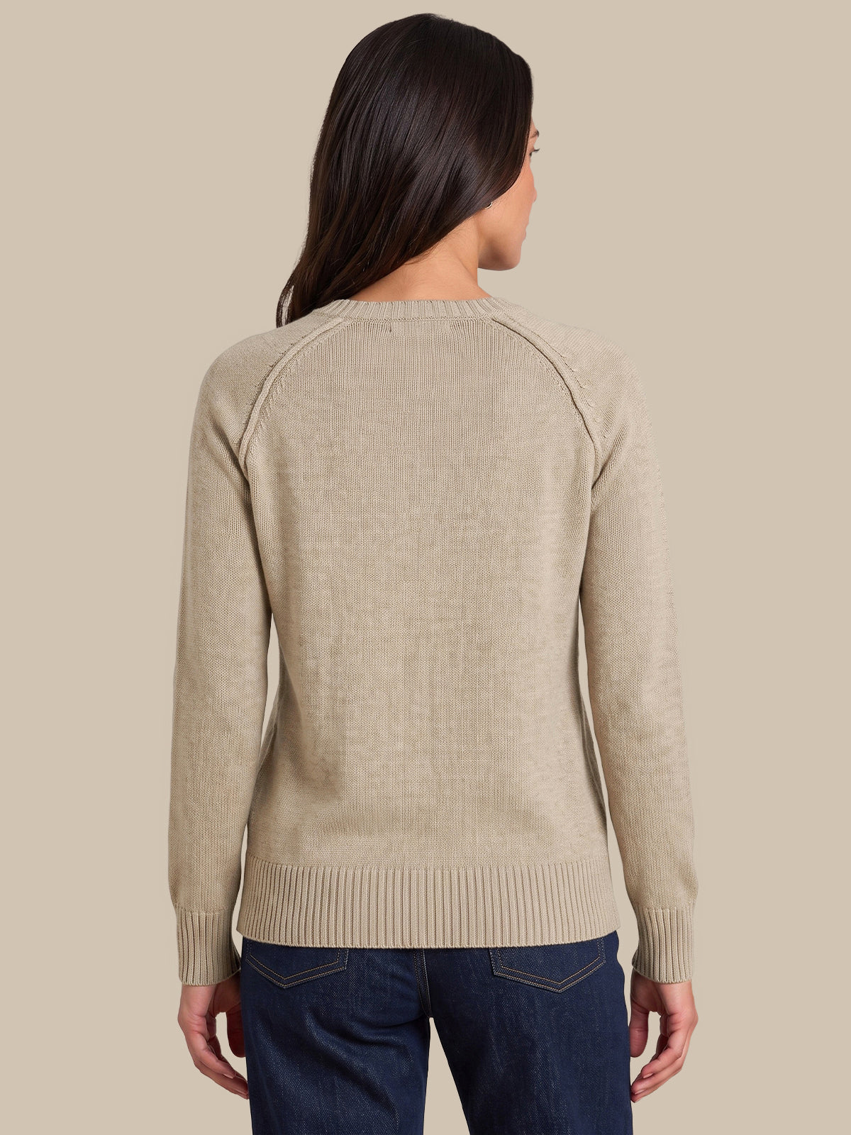 A woman viewed from the back wearing a light beige knit sweater with raglan sleeves and dark blue jeans, set against a solid beige background.
