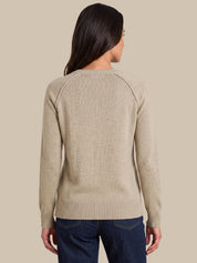 A woman viewed from the back wearing a light beige knit sweater with raglan sleeves and dark blue jeans, set against a solid beige background.