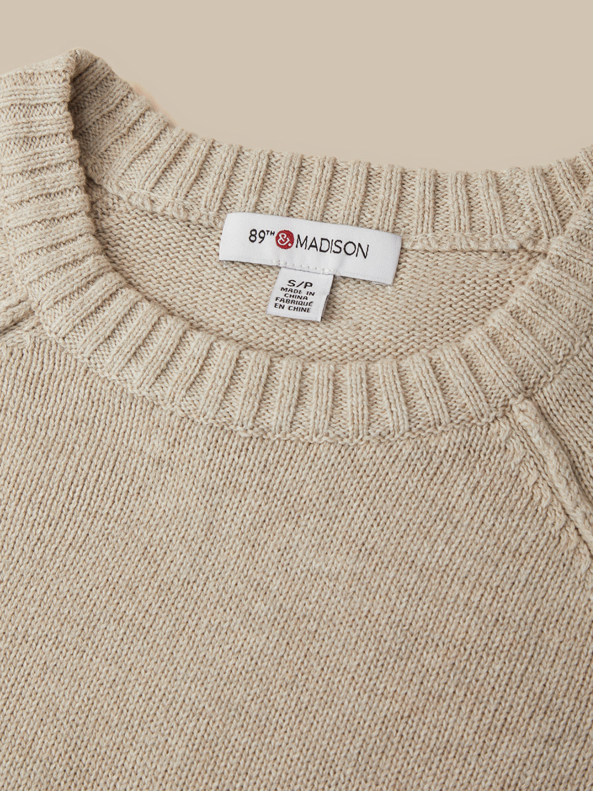 Close-up of a beige knit sweater with a ribbed crew neck and a visible neck label reading 89th & Madison.