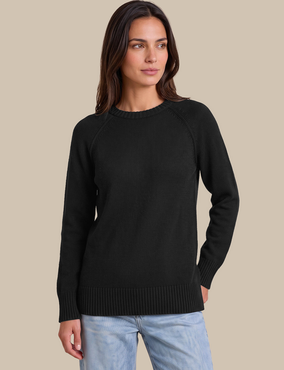 A woman wearing a black long-sleeve knit sweater with raglan sleeves and ribbed trim, paired with light blue jeans, set against a solid beige background.