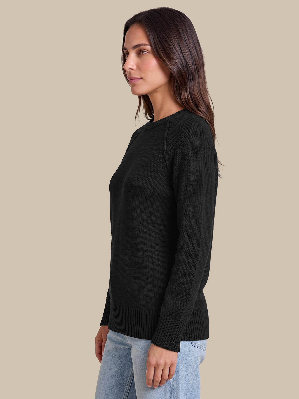 A woman shown in side profile wearing a black long-sleeve knit sweater and light blue jeans against a plain beige studio background.