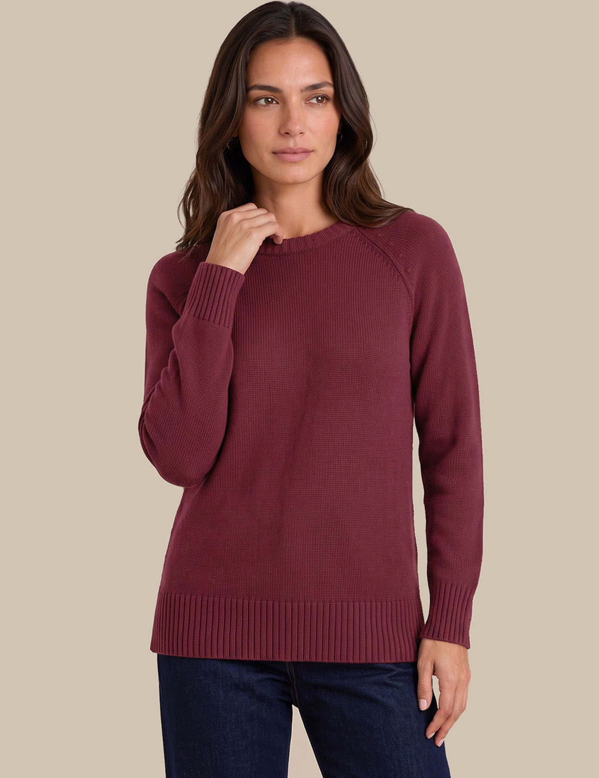 A woman modeling a long-sleeve crewneck sweater in burgundy, paired with dark blue jeans, against a solid beige background.