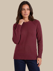 A woman modeling a long-sleeve crewneck sweater in burgundy, paired with dark blue jeans, against a solid beige background.