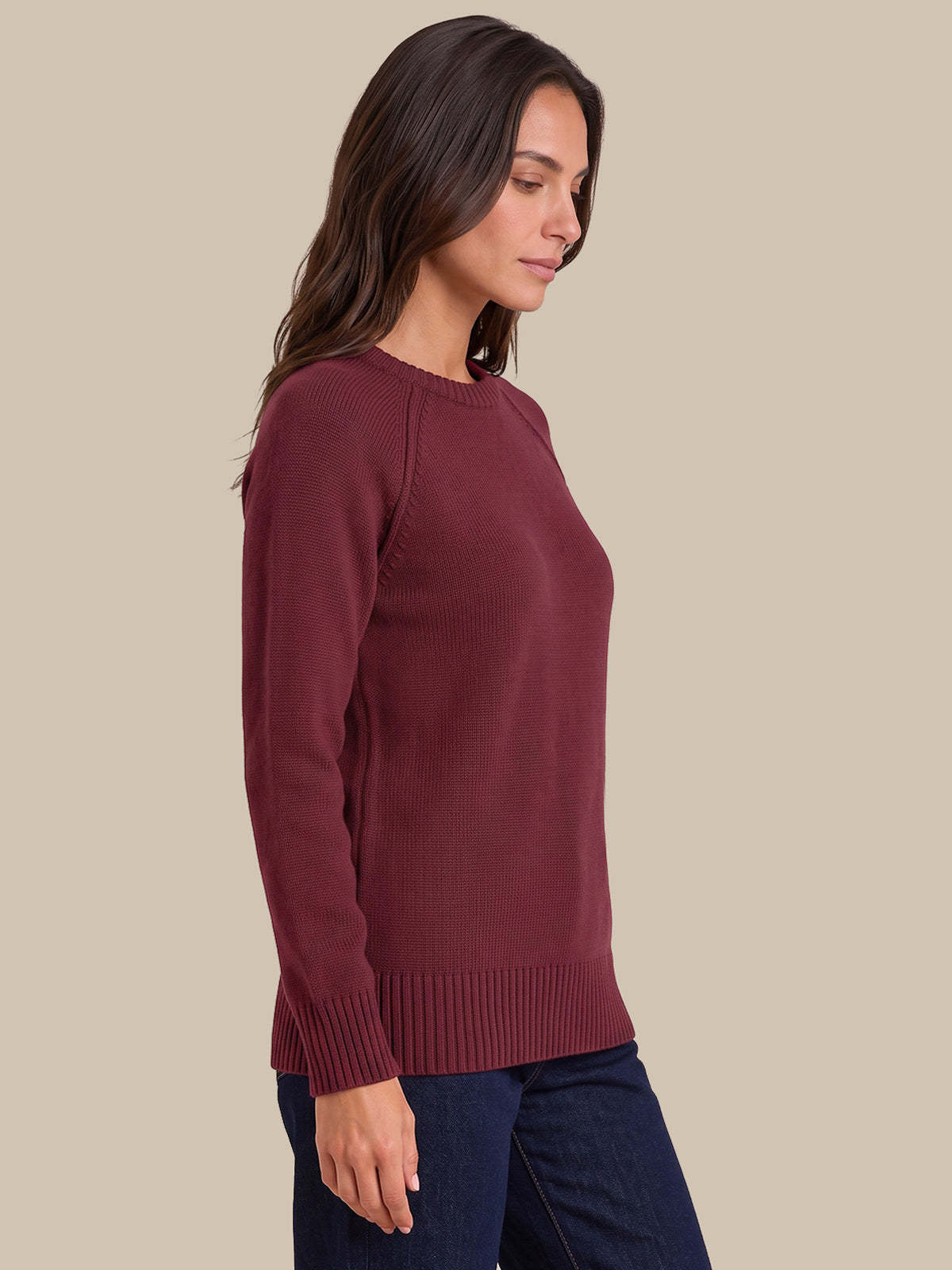 A woman wearing a long-sleeve crewneck knit sweater in burgundy, paired with dark blue jeans, shown in a side-profile view against a solid beige background.