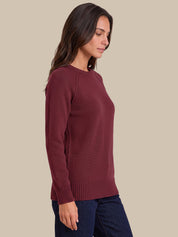 A woman wearing a long-sleeve crewneck knit sweater in burgundy, paired with dark blue jeans, shown in a side-profile view against a solid beige background.