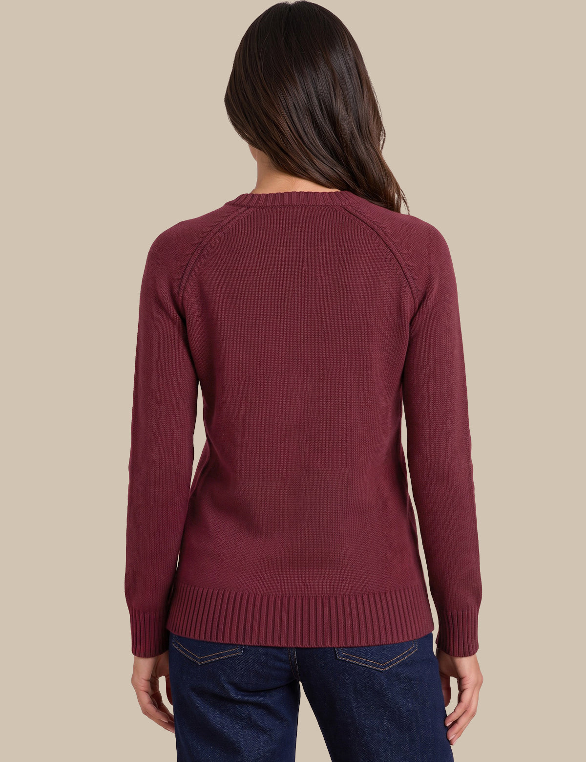 A woman viewed from the back wearing a long-sleeve ribbed knit sweater in a deep burgundy color, paired with dark blue jeans, against a solid beige background.