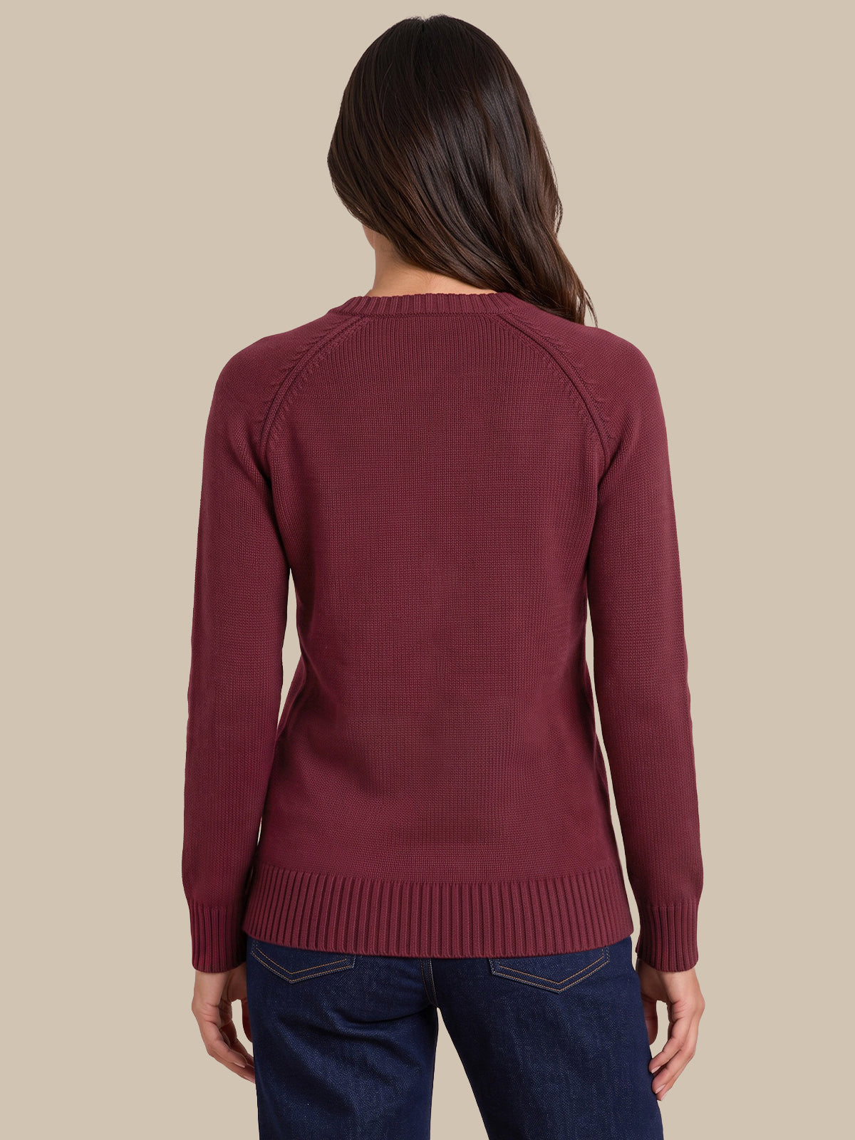 A woman viewed from the back wearing a long-sleeve ribbed knit sweater in a deep burgundy color, paired with dark blue jeans, against a solid beige background.