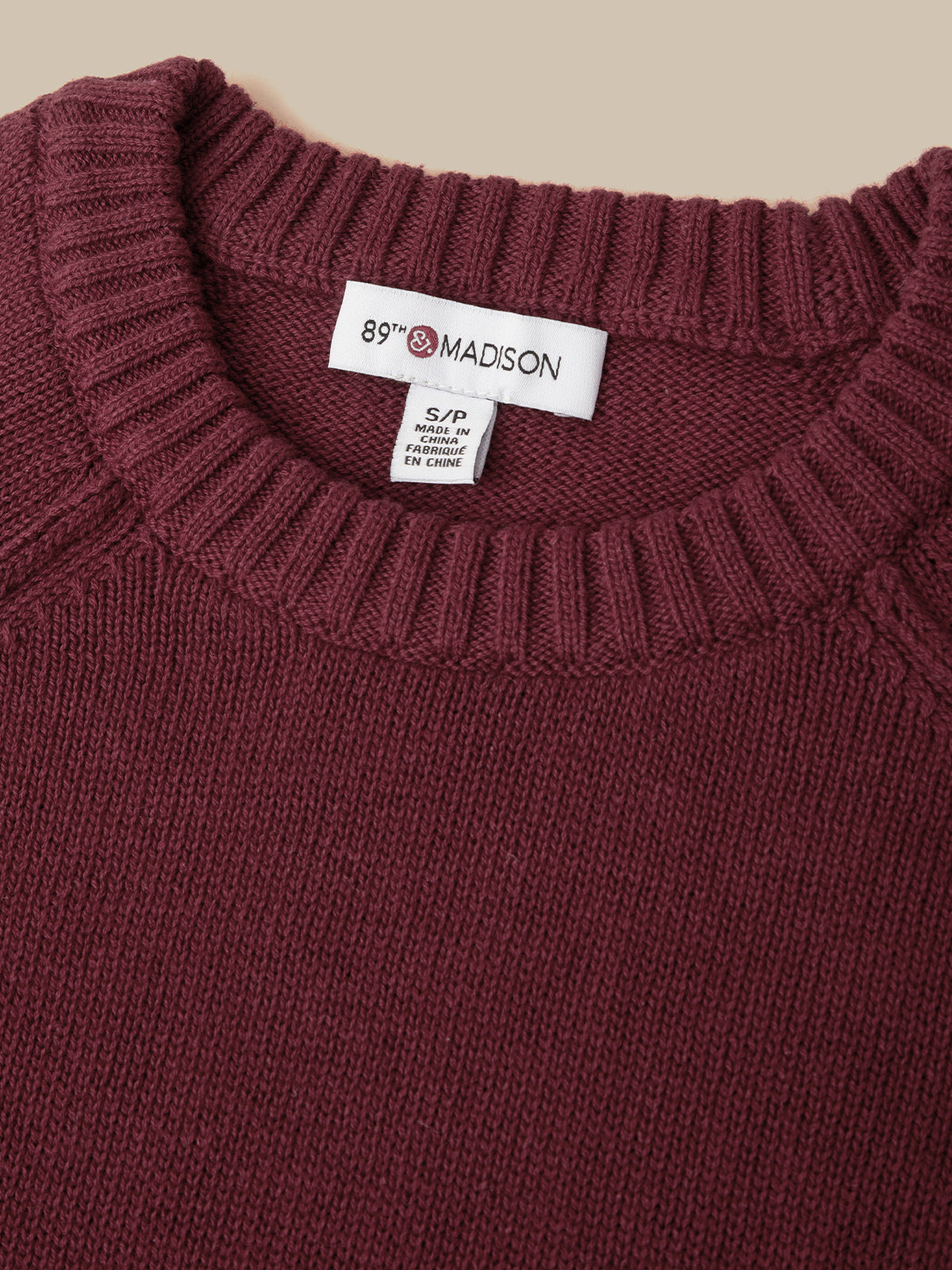 Close-up of a ribbed knit crewneck sweater in deep burgundy, showing the white 89th Madison brand label at the collar.