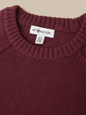 Close-up of a ribbed knit crewneck sweater in deep burgundy, showing the white 89th Madison brand label at the collar.