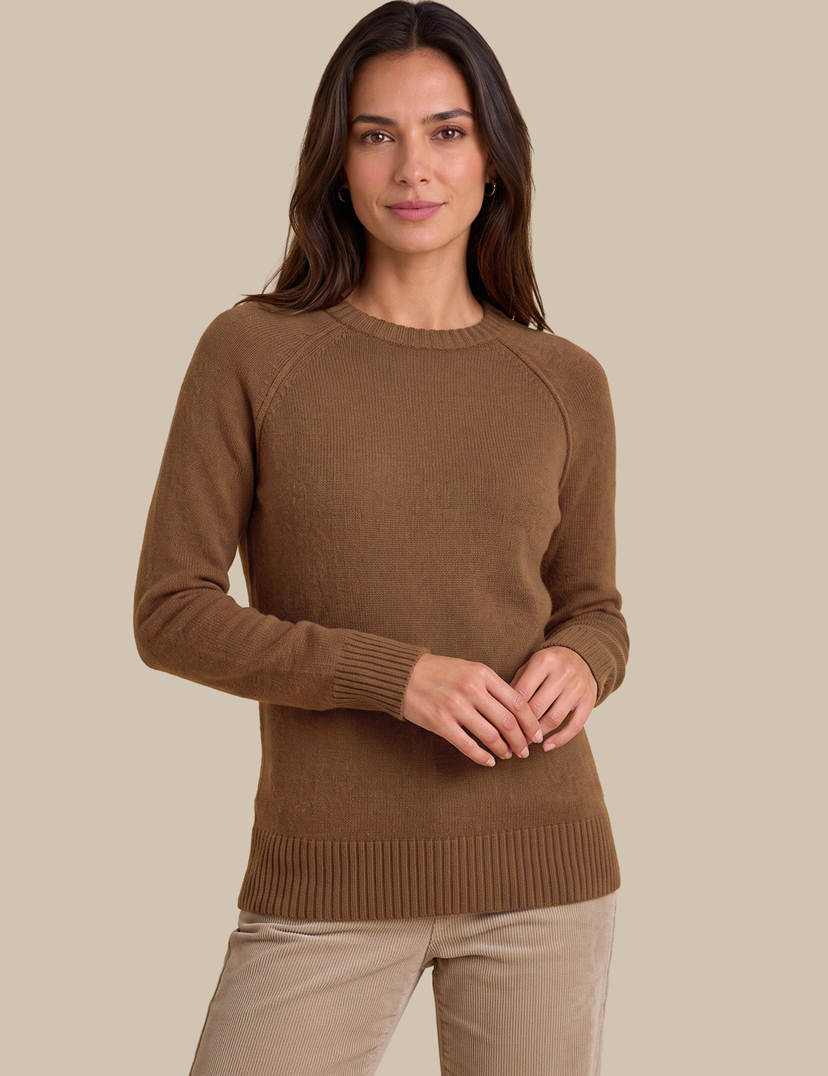 A woman wearing a brown long-sleeve knit sweater and beige pants, standing against a solid beige studio background.