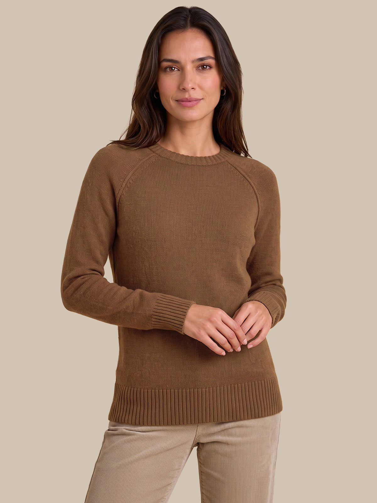 A woman wearing a brown long-sleeve knit sweater and beige pants, standing against a solid beige studio background.