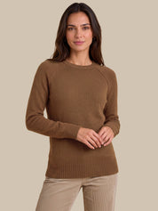 A woman wearing a brown long-sleeve knit sweater and beige pants, standing against a solid beige studio background.