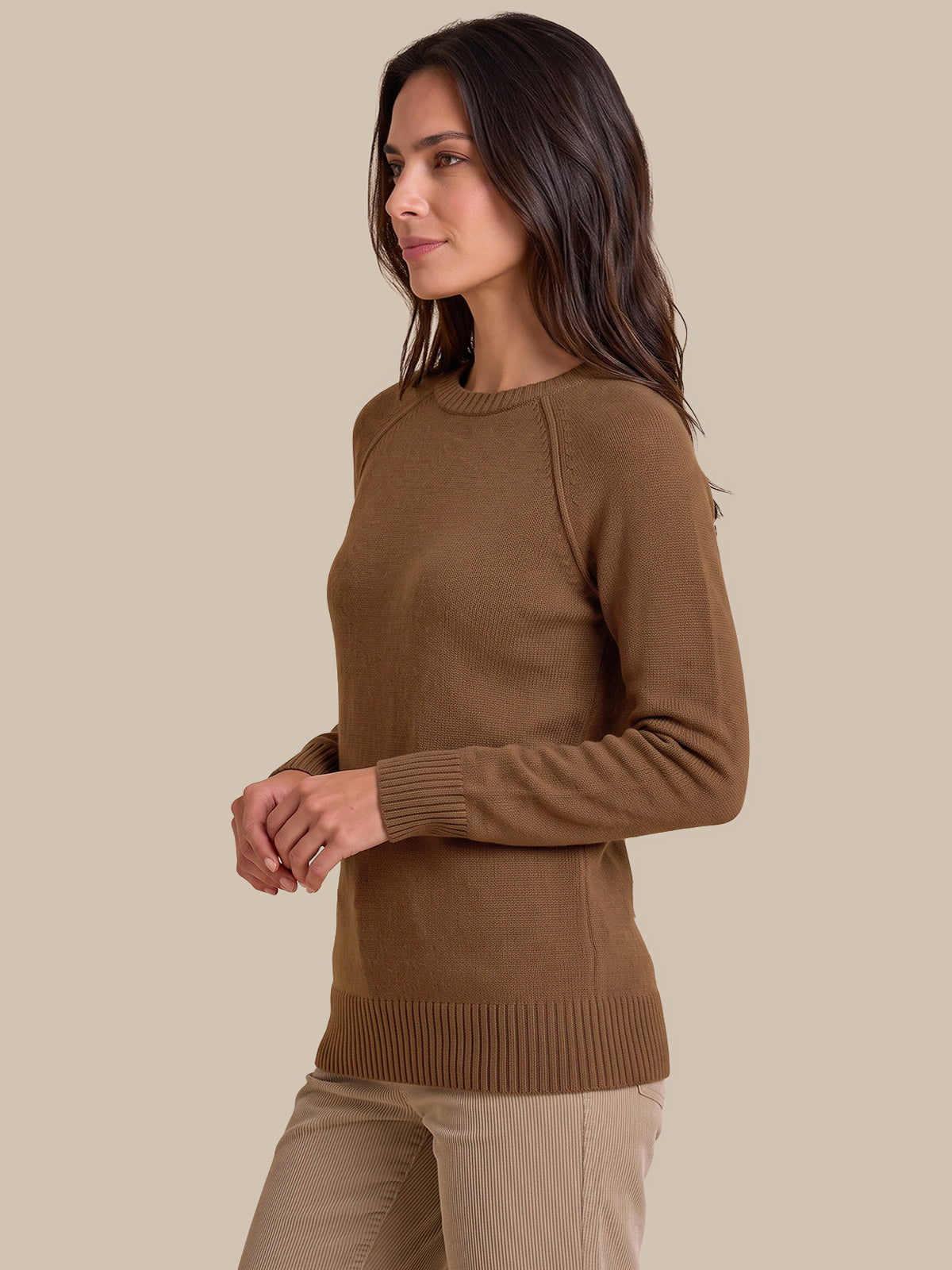 A woman wearing a brown long-sleeve knit sweater and beige pants, shown in profile against a plain beige studio background.