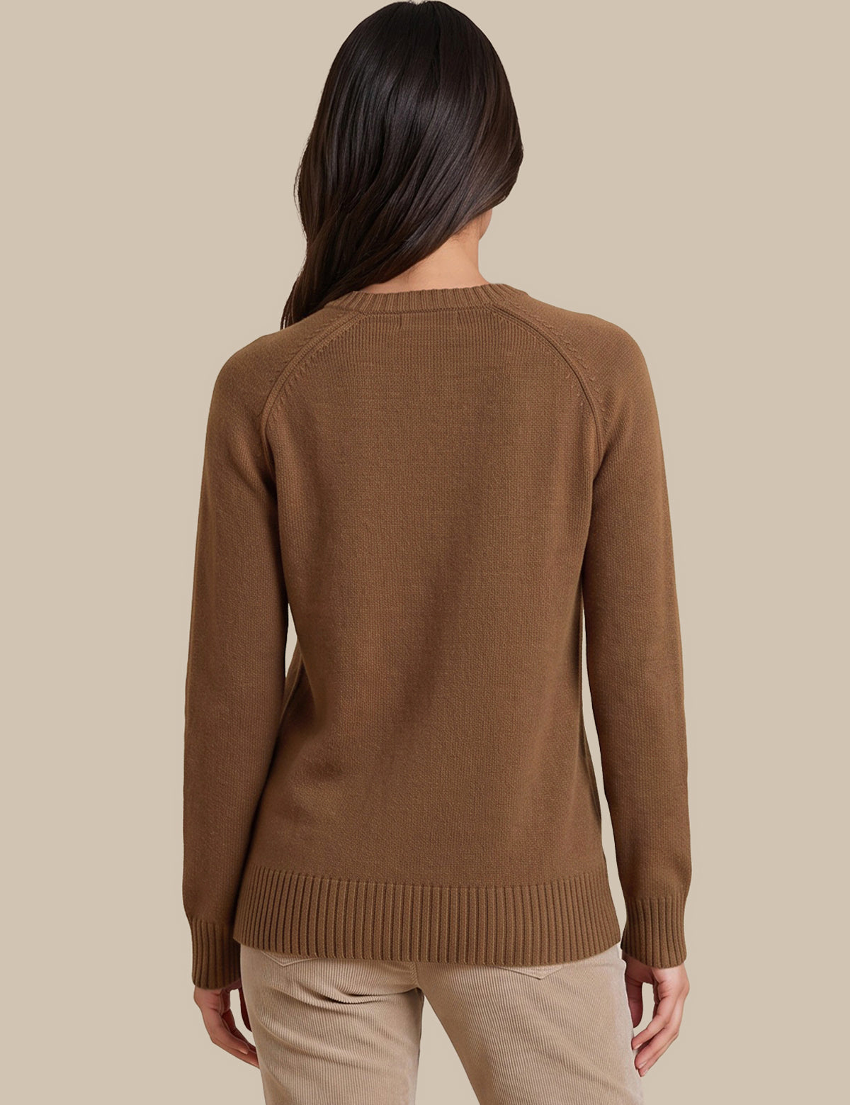 A woman viewed from the back wearing a brown ribbed knit sweater and beige pants, set against a solid beige studio background.