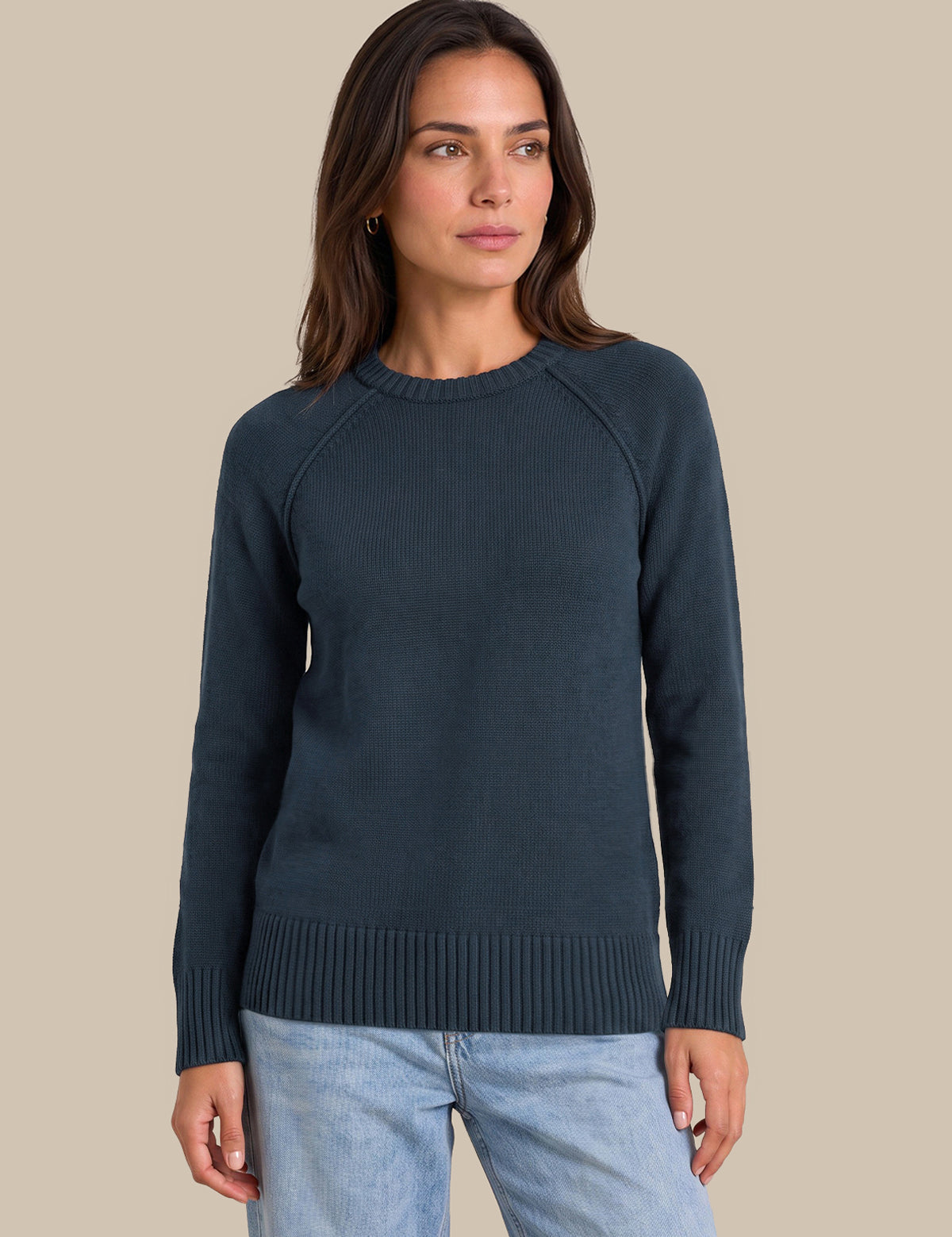 A woman wearing a dark slate blue ribbed knit sweater and light blue jeans, standing against a solid beige background.