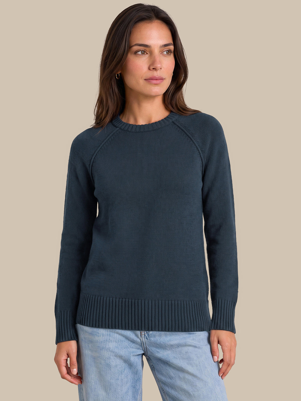 A woman wearing a dark slate blue ribbed knit sweater and light blue jeans, standing against a solid beige background.