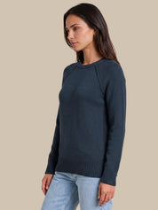 A woman wearing a dark slate blue crewneck knit sweater and light blue jeans, shown from a side profile against a solid beige background.