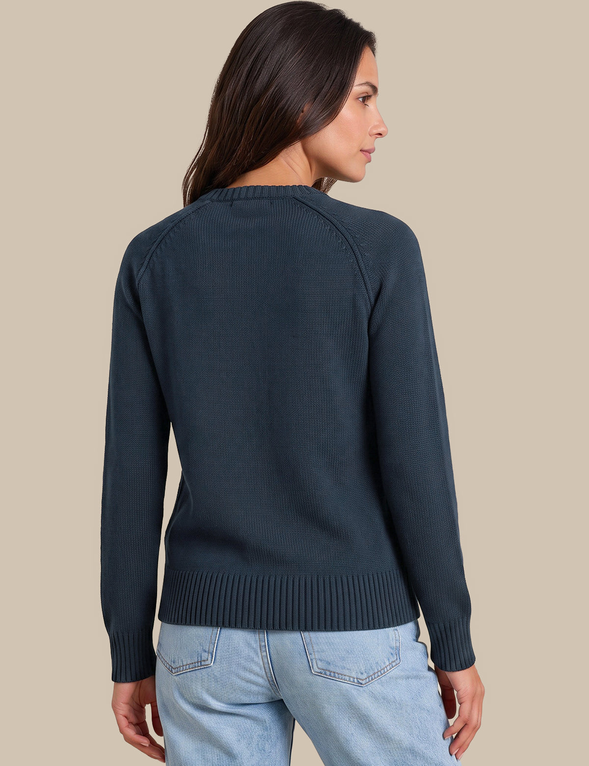 A woman shown from the back wearing a dark navy blue knit sweater and light blue jeans, set against a solid beige background.