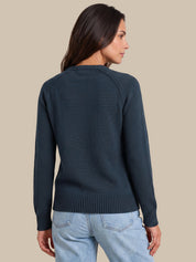 A woman shown from the back wearing a dark navy blue knit sweater and light blue jeans, set against a solid beige background.