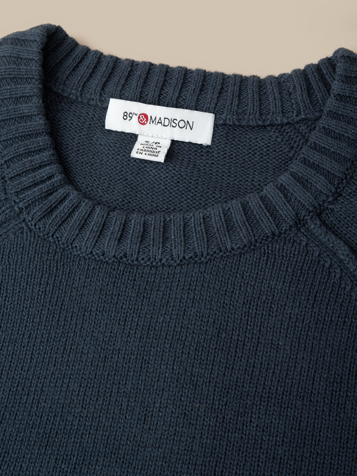 Close-up of a dark navy blue knit sweater with a ribbed crew neck, featuring a white neck label with the brand name '89° MADISON'.