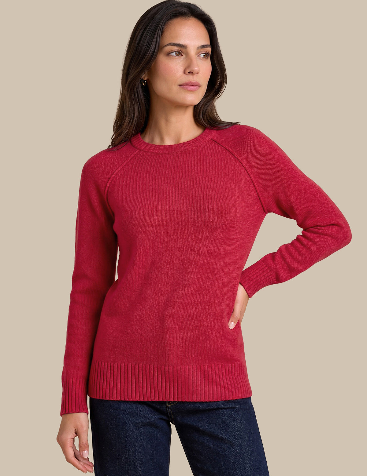 A woman modeling a long-sleeve crewneck knit sweater in a deep red color, paired with dark blue jeans, against a solid beige background.