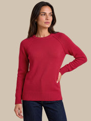 A woman modeling a long-sleeve crewneck knit sweater in a deep red color, paired with dark blue jeans, against a solid beige background.
