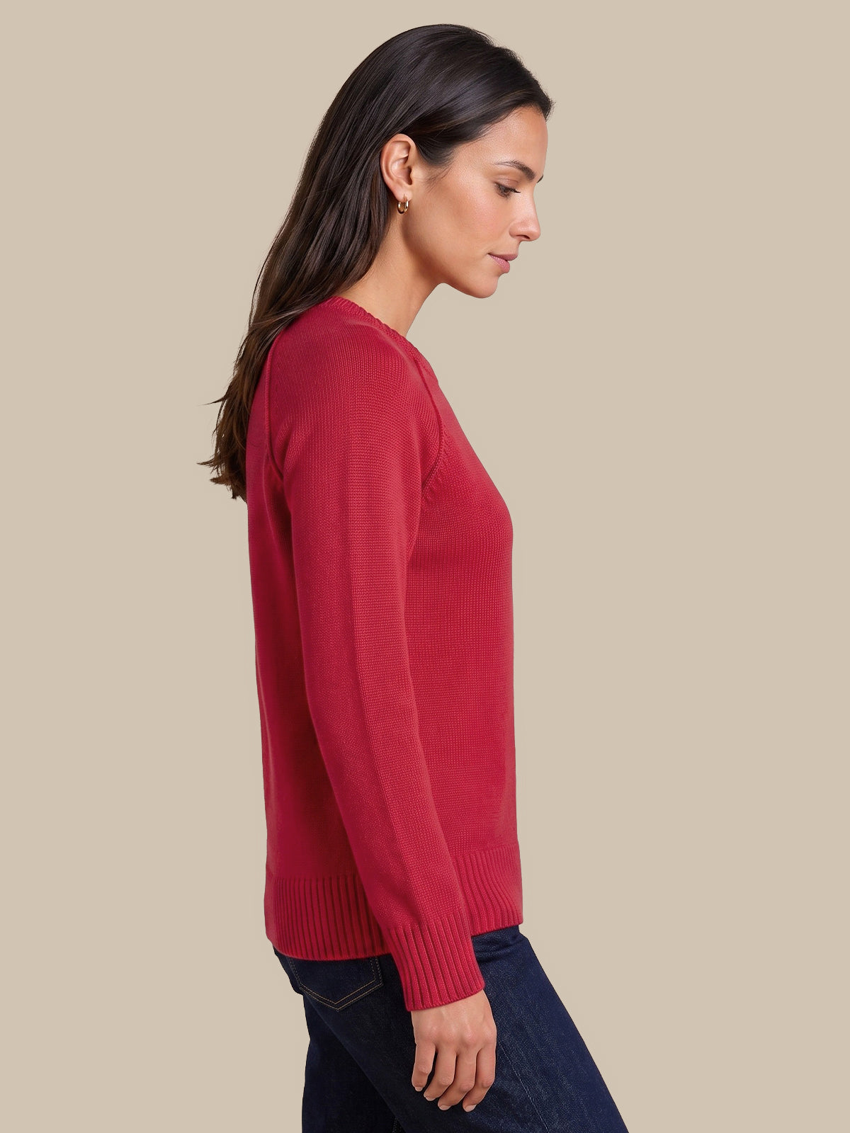 A woman in side profile wearing a solid red long-sleeve knit sweater and dark blue jeans, set against a solid beige studio background.