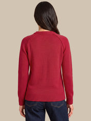 A woman viewed from the back wearing a red knit sweater and dark blue jeans against a solid beige background.