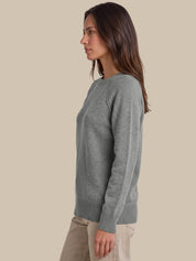 A woman shown in side profile wearing a light gray knit sweater and beige pants against a solid beige studio background.