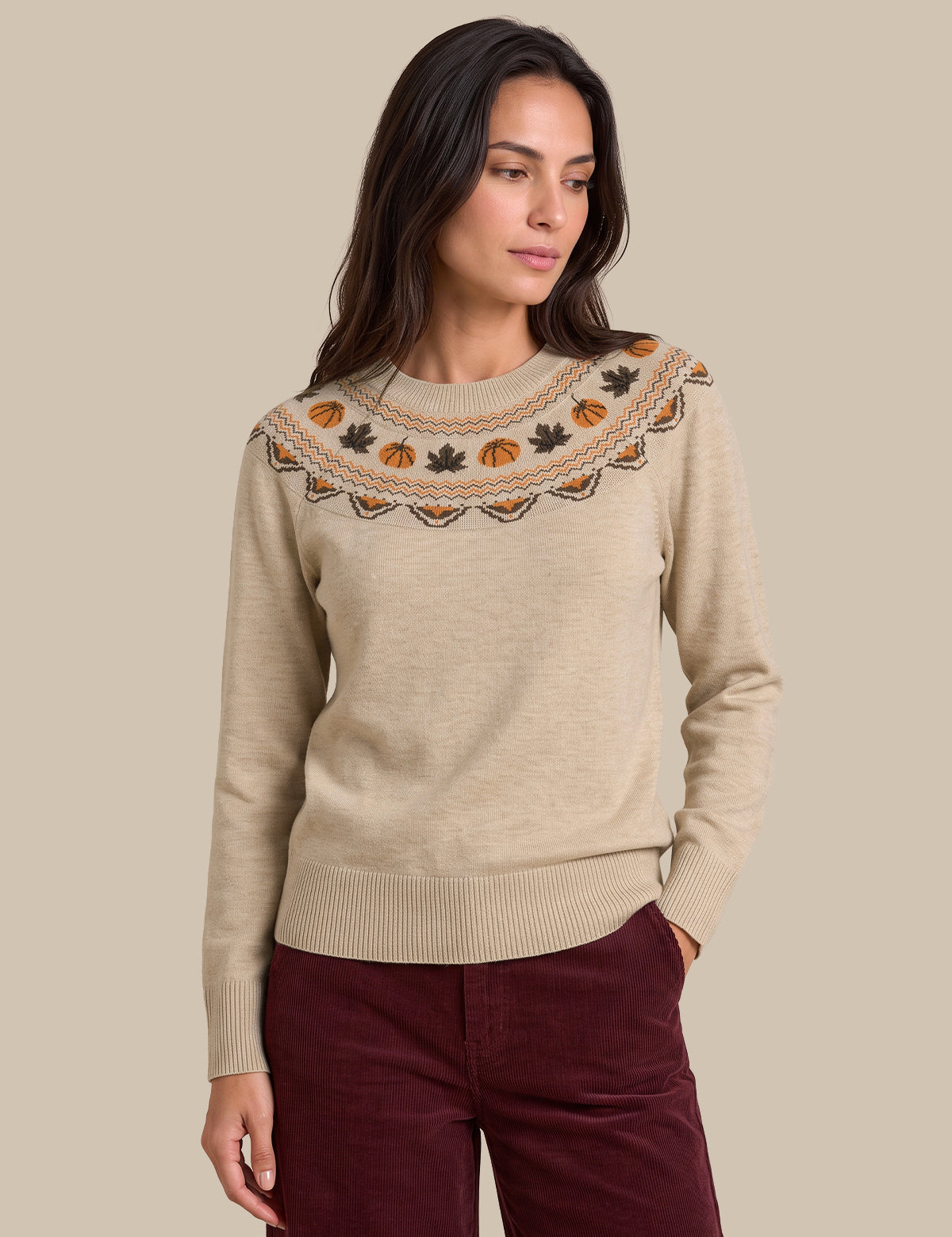 A woman modeling a beige knit sweater with a decorative embroidered yoke featuring autumn motifs like pumpkins, leaves, and pinecones in orange, brown, and black, paired with burgundy pants against a solid beige background.