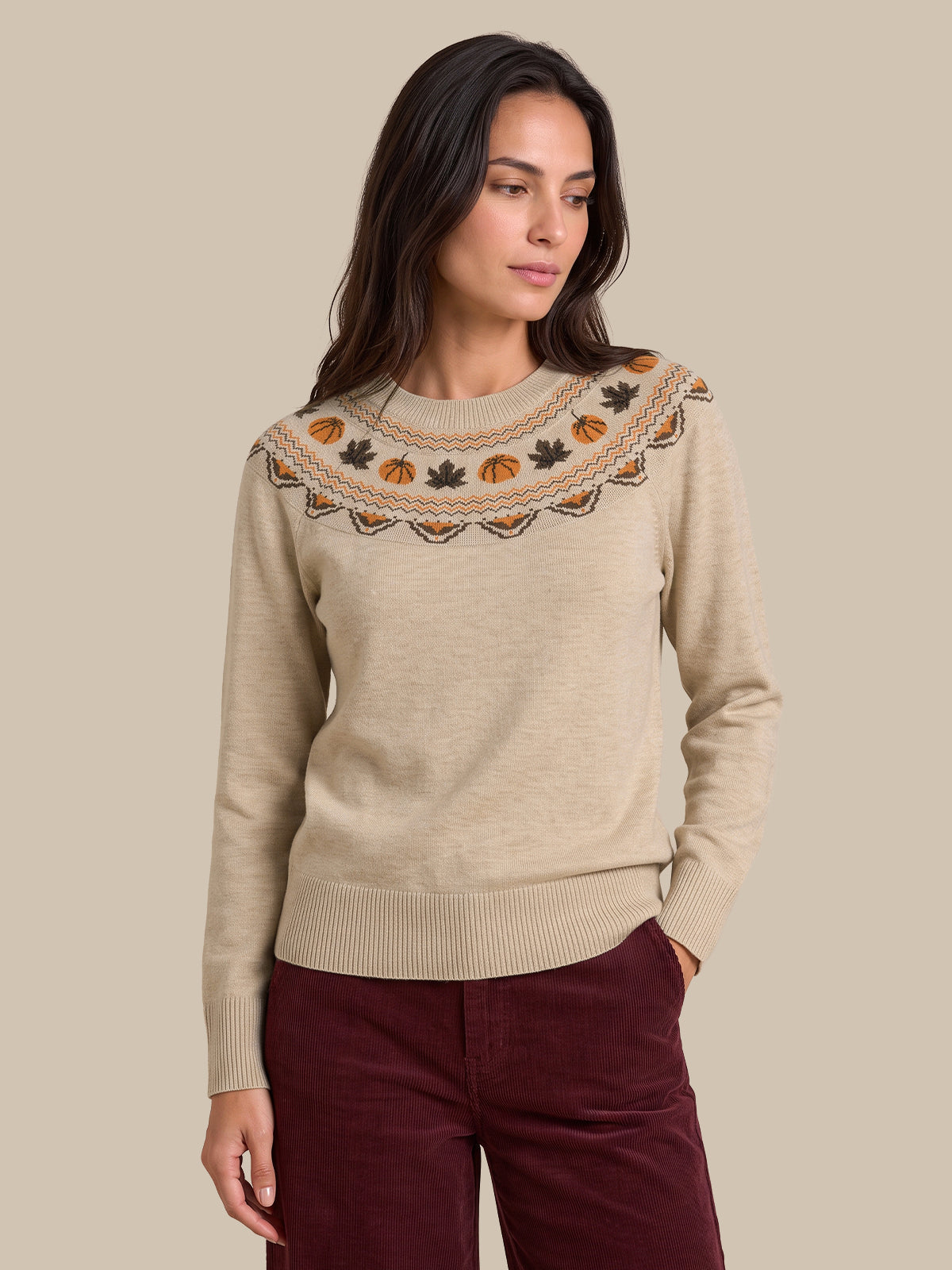 A woman modeling a beige knit sweater with a decorative embroidered yoke featuring autumn motifs like pumpkins, leaves, and pinecones in orange, brown, and black, paired with burgundy pants against a solid beige background.