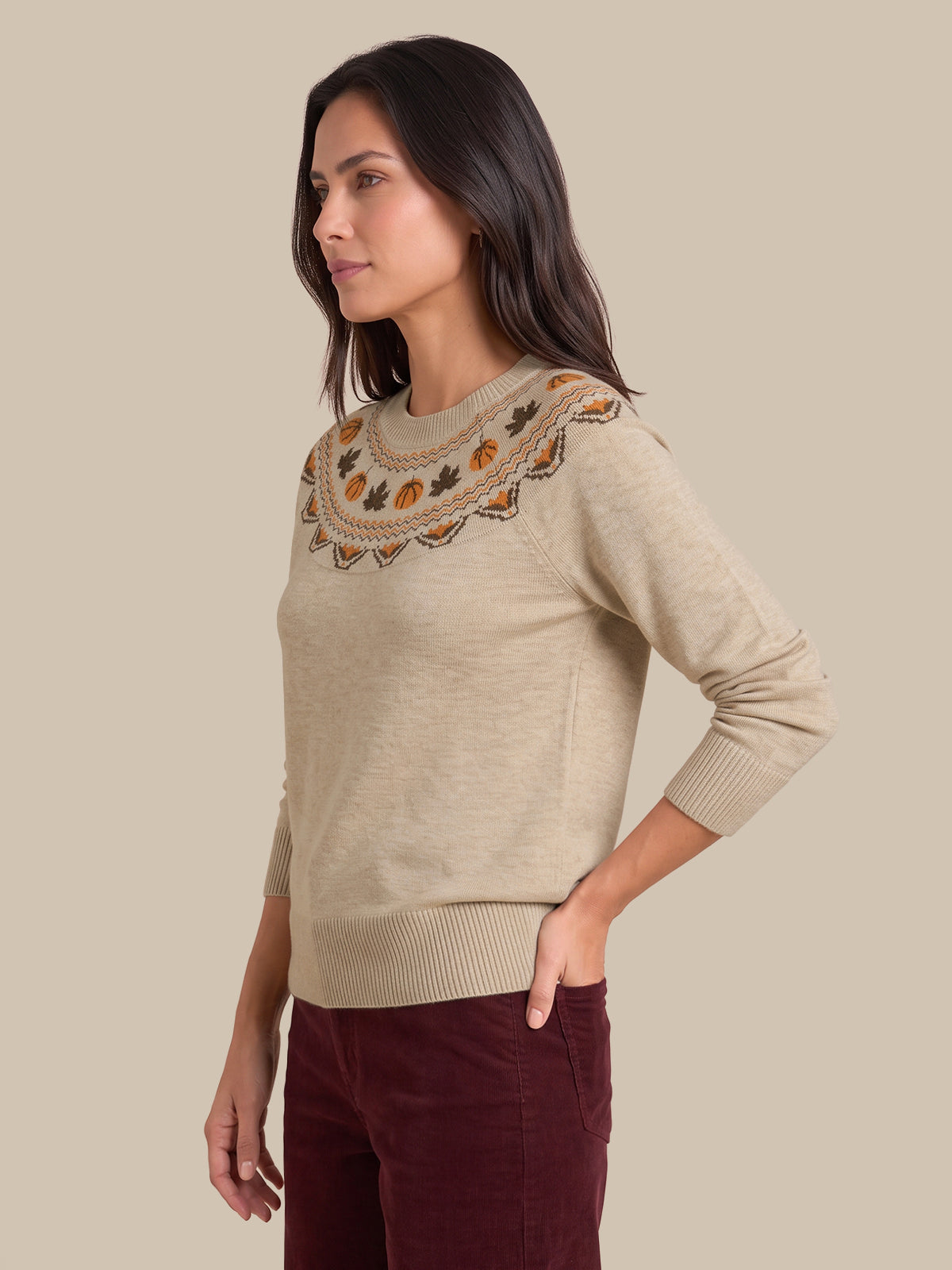 A woman wearing a beige knit sweater with a decorative embroidered yoke in orange, brown, and black, paired with burgundy pants, set against a solid beige background.