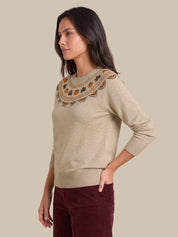 A woman wearing a beige knit sweater with a decorative embroidered yoke in orange, brown, and black, paired with burgundy pants, set against a solid beige background.