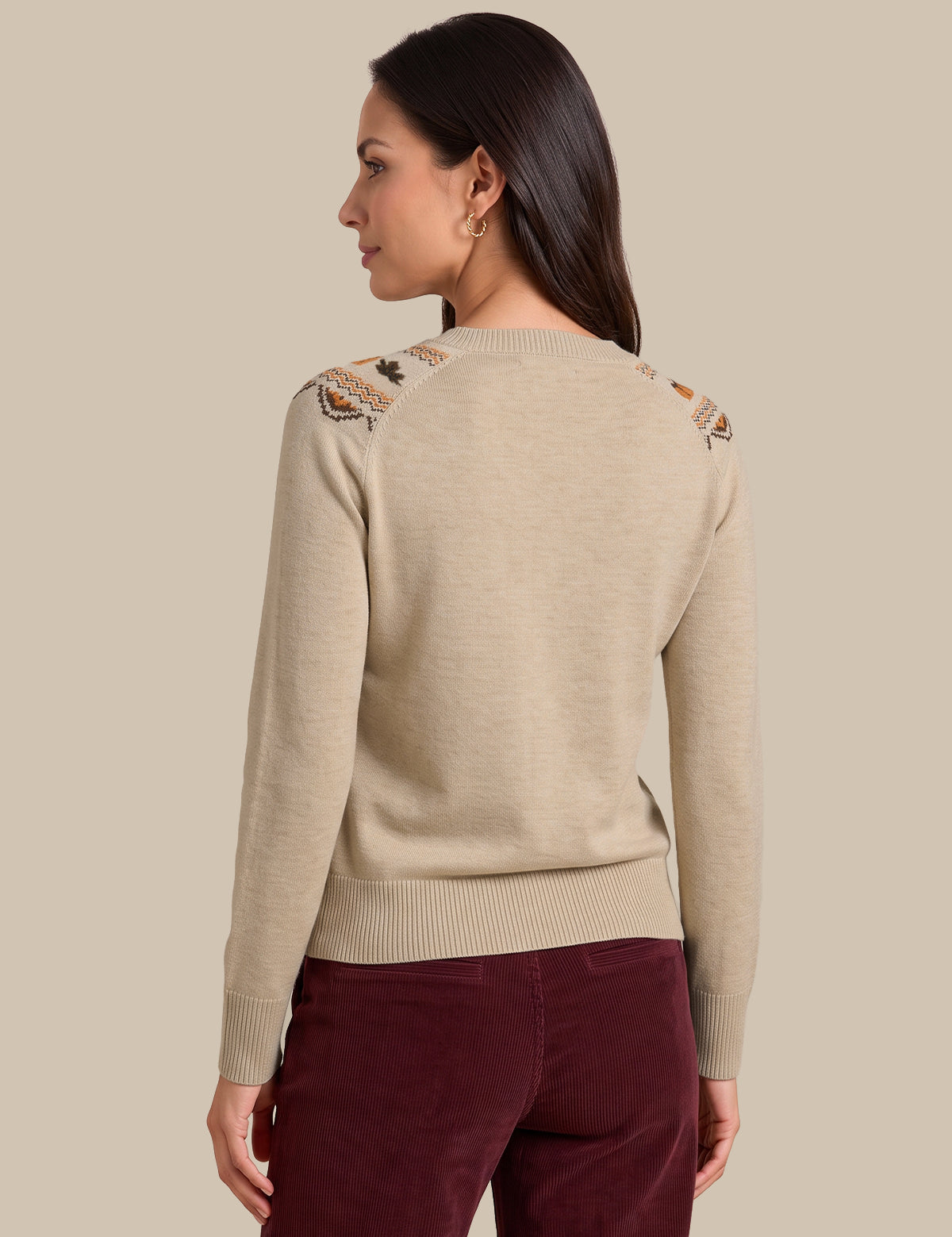 A woman shown from the back wearing a light beige knit sweater with decorative embroidered details on the shoulders and burgundy pants, set against a solid beige background.