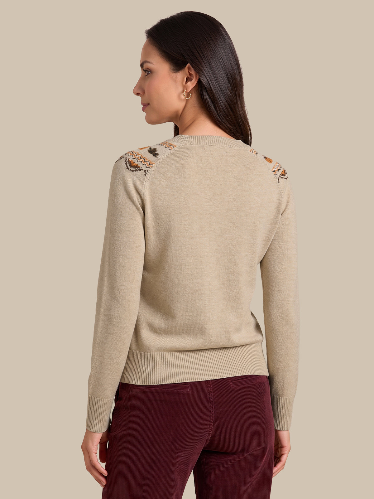 A woman shown from the back wearing a light beige knit sweater with decorative embroidered details on the shoulders and burgundy pants, set against a solid beige background.