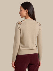 A woman shown from the back wearing a light beige knit sweater with decorative embroidered details on the shoulders and burgundy pants, set against a solid beige background.