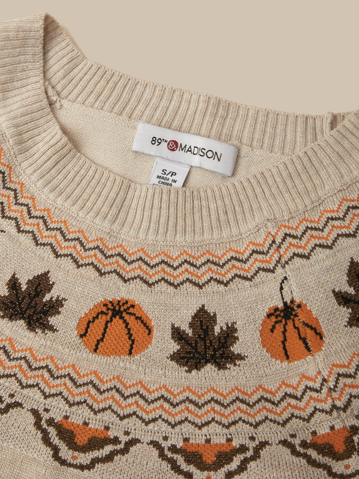 Close-up of a cream knit sweater with a pattern of orange pumpkins and dark brown maple leaves, featuring a white neck label that reads 89th & MADISON.