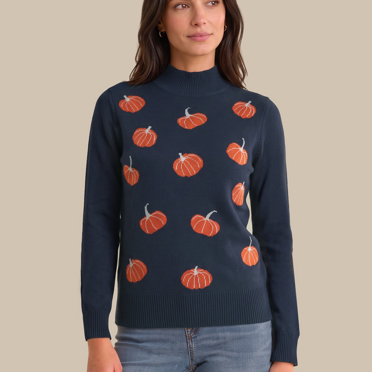 A woman wearing a navy blue mock neck sweater with an orange pumpkin pattern, paired with light blue jeans, against a solid beige background.