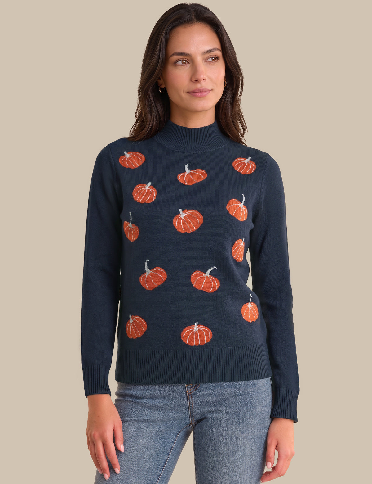 A woman wearing a navy blue mock neck sweater with an orange pumpkin pattern, paired with light blue jeans, against a solid beige background.