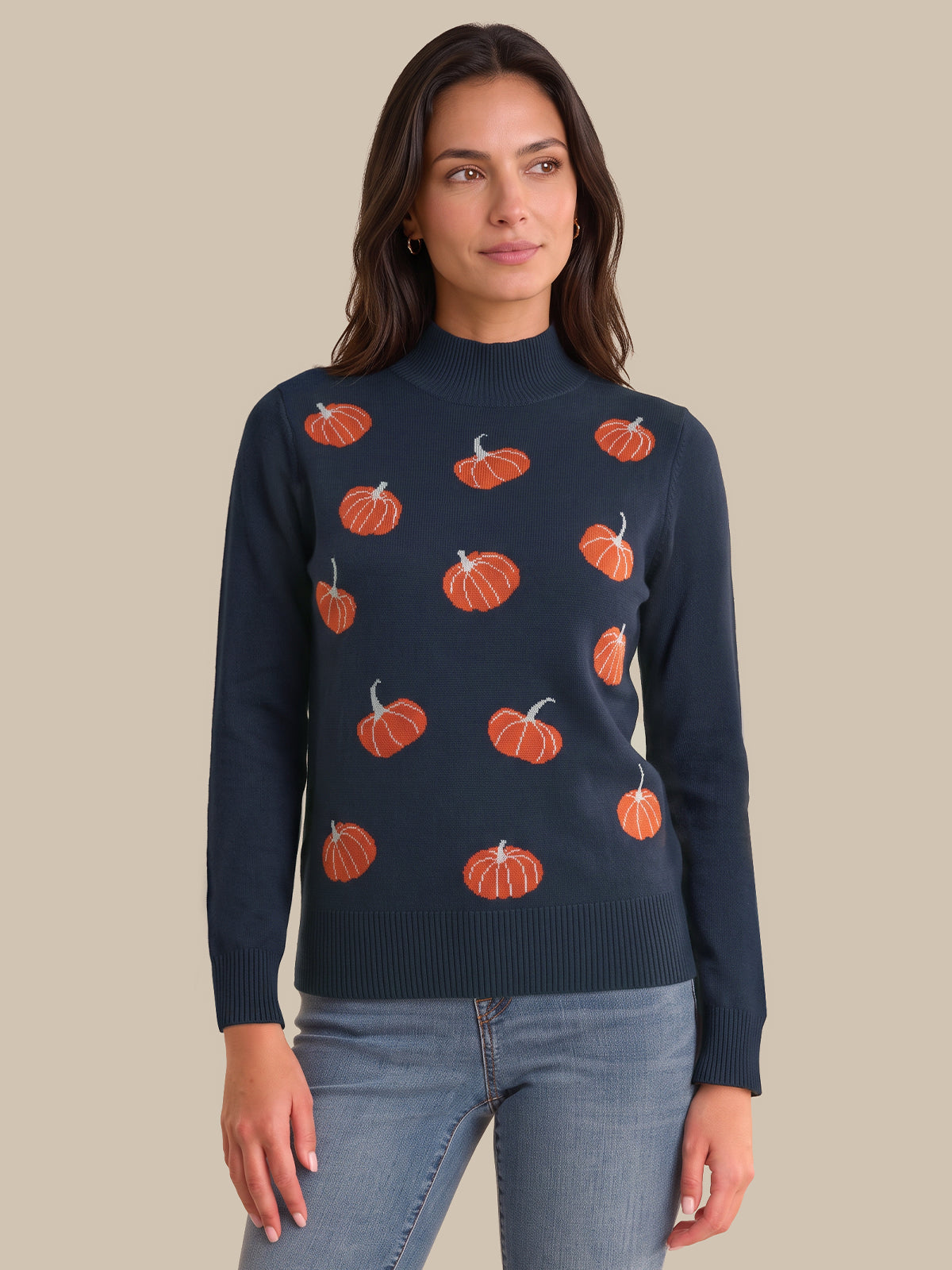 A woman wearing a navy blue mock neck sweater with an orange pumpkin pattern, paired with light blue jeans, against a solid beige background.
