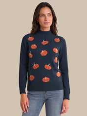A woman wearing a navy blue mock neck sweater with an orange pumpkin pattern, paired with light blue jeans, against a solid beige background.