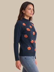 A woman wearing a navy blue knit sweater with an orange pumpkin pattern, paired with light blue jeans, shown against a solid beige background.