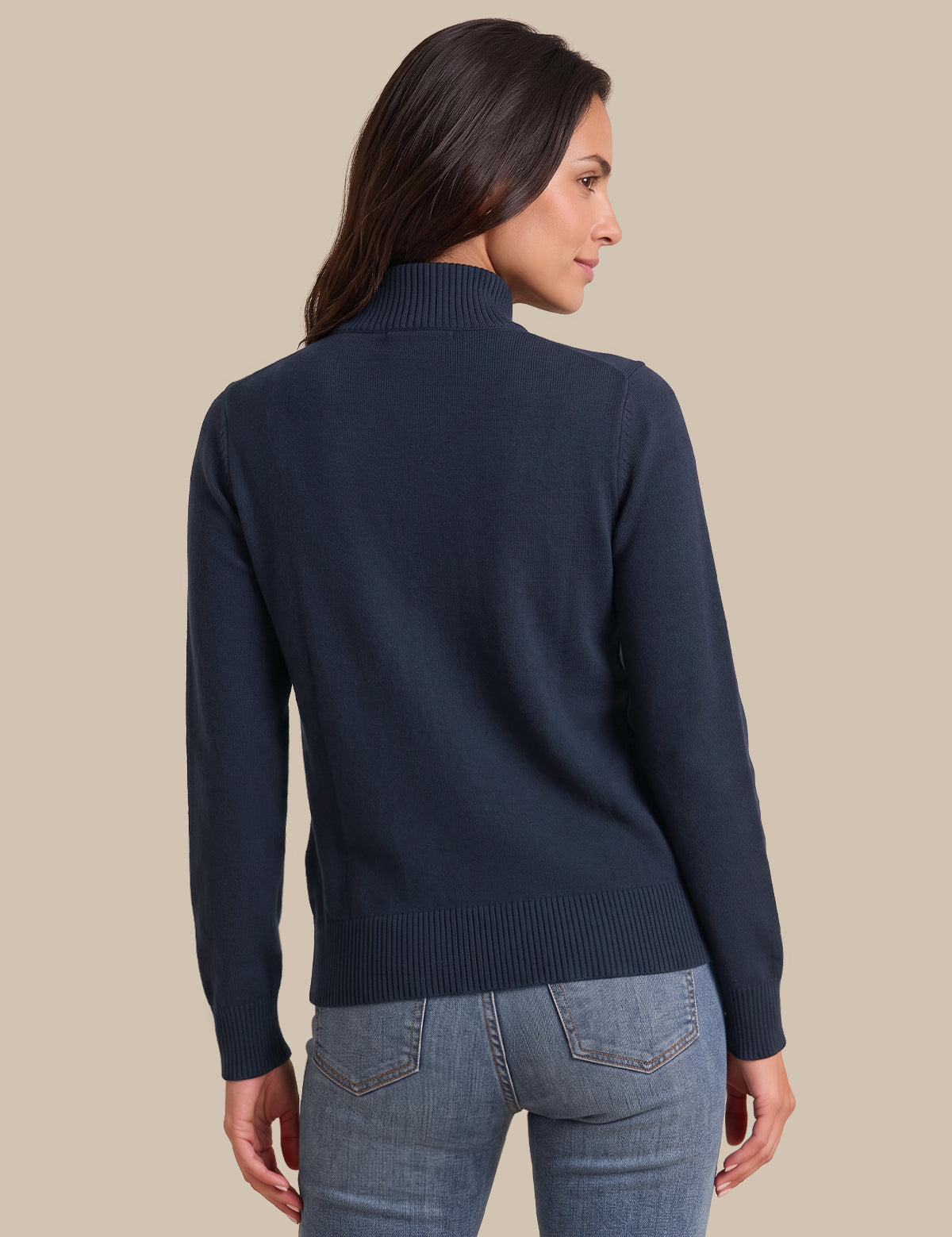 A woman shown from the back wearing a navy blue mock neck knit sweater and light blue jeans, set against a solid beige background.