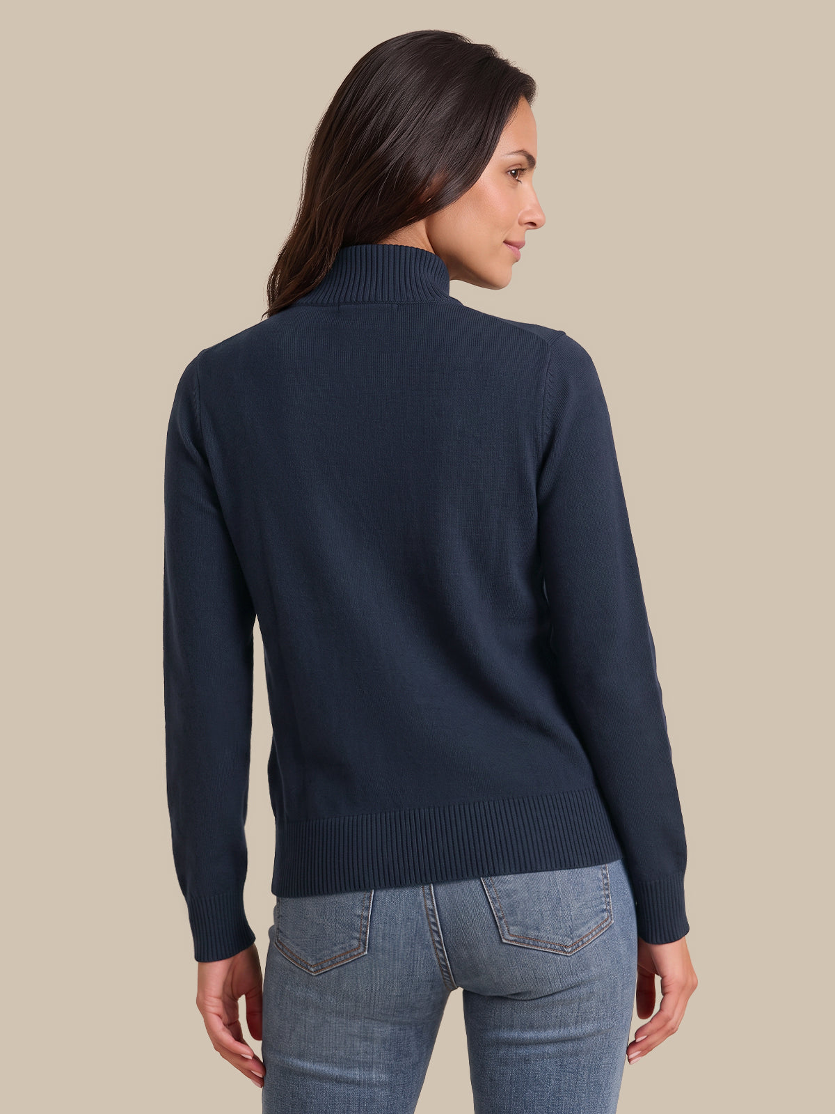 A woman shown from the back wearing a navy blue mock neck knit sweater and light blue jeans, set against a solid beige background.
