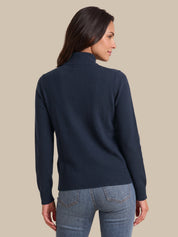 A woman shown from the back wearing a navy blue mock neck knit sweater and light blue jeans, set against a solid beige background.