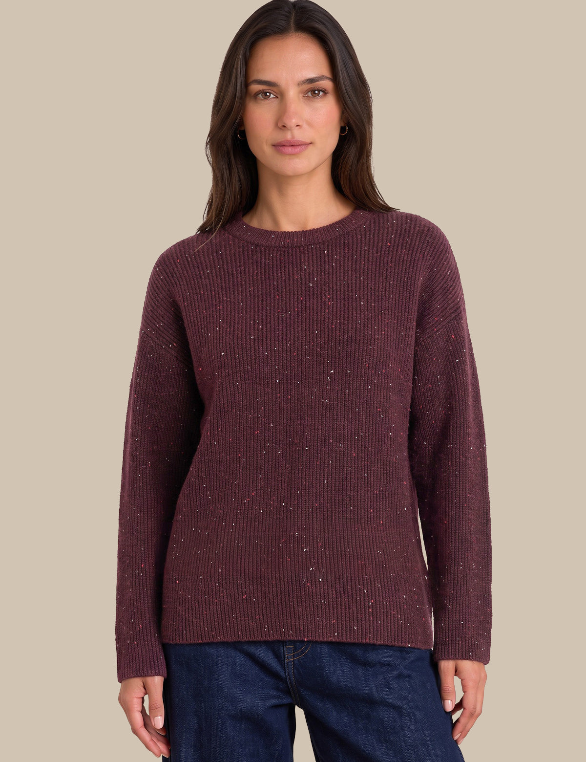 A woman wearing a ribbed long-sleeve knit sweater in a deep burgundy color with subtle silver flecks, paired with dark blue jeans, set against a solid beige background.