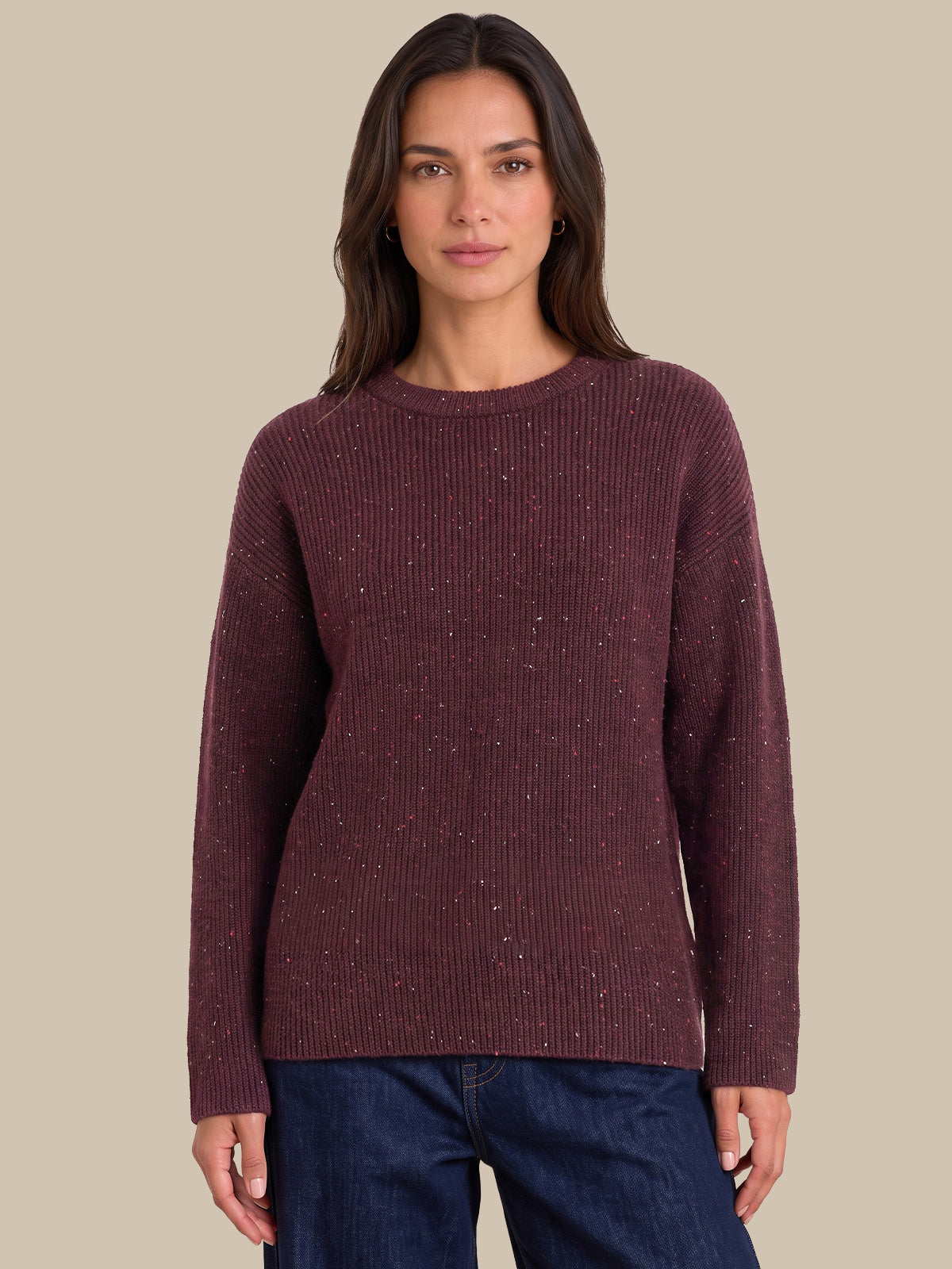 A woman wearing a ribbed long-sleeve knit sweater in a deep burgundy color with subtle silver flecks, paired with dark blue jeans, set against a solid beige background.