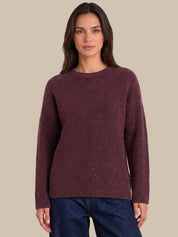 A woman wearing a ribbed long-sleeve knit sweater in a deep burgundy color with subtle silver flecks, paired with dark blue jeans, set against a solid beige background.