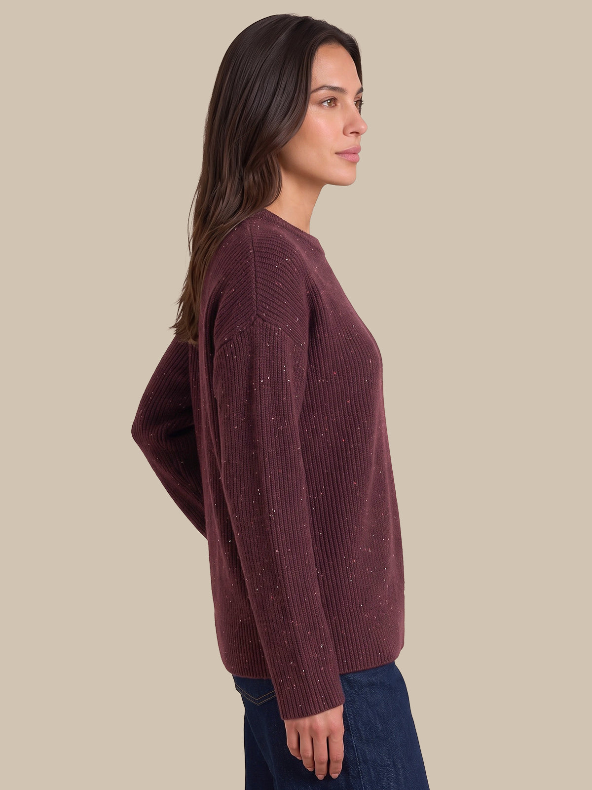 A woman shown in side profile wearing a ribbed knit sweater in a deep plum-burgundy color with subtle white flecks, paired with dark blue jeans, against a solid beige background.